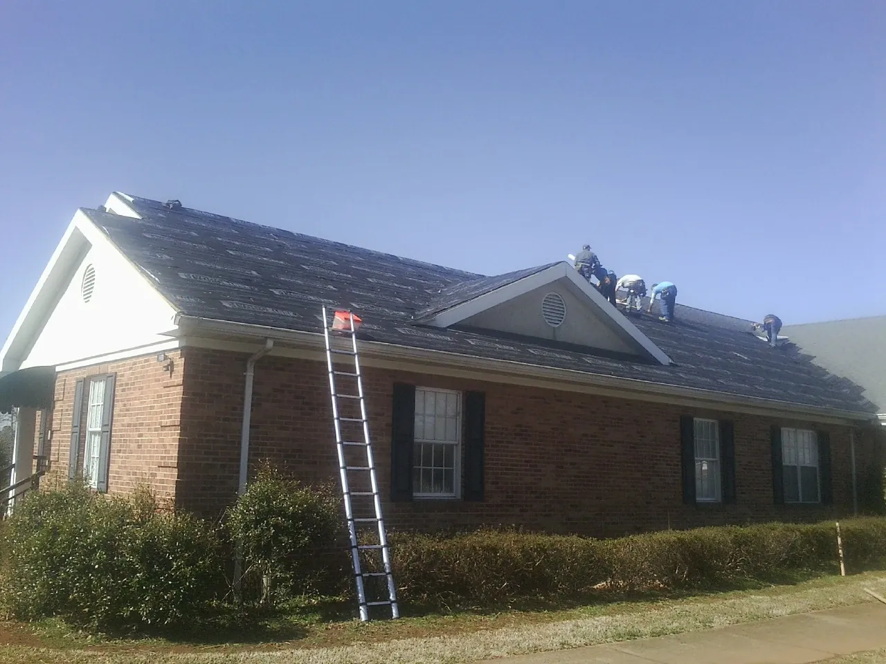 Crew tearing shingles off a brick ranch roof with a ladder set — Emergency Roof Repair work by our crew in Ross